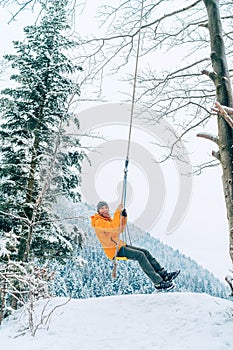 A young man in an orange softshell jacket laughing while he swinging on a swing between forest trees with picturesque snowy