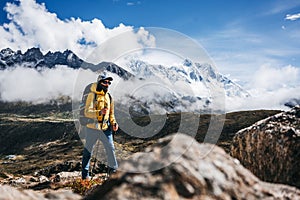 Young man outdoor traveler walk along huge sunny cloudy mountains way. Tourist with backpack and trekking poles going across