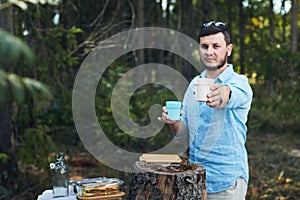 A young man at a picnic holds out a glass with a drink to the camera.