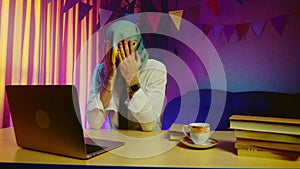 A young man in a pigeon mask is sitting on a chair at a table and typing on a laptop keyboard. This man is desperate