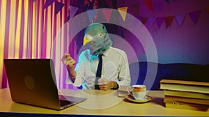 A young man in a pigeon mask is sitting on a chair at a table and typing on a laptop keyboard. The man is angry