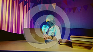 A young man in a pigeon mask is sitting on a chair at a table typing on a laptop keyboard. Multicolored lighting