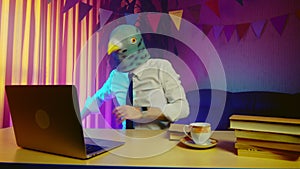 A young man in a pigeon mask is sitting on a chair at a table and typing on a laptop keyboard. This person is happy