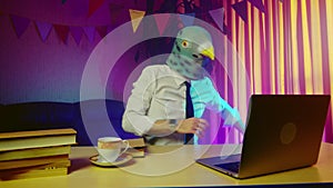 A young man in a pigeon mask is sitting on a chair at a table and typing on a laptop keyboard. This person is happy