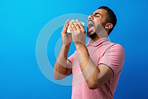 Young man in pink shirt with allergy or cold, blowing his nose in a tissue against blue background