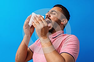 Young man in pink shirt with allergy or cold, blowing his nose in a tissue against blue background