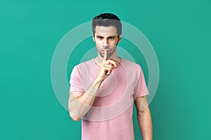Young man in pink t-shirt keeps finger on lips, making hush gesture isolated on blue background