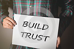 Young man in plaid shirt holding a placard with text: BUILD TRUST!