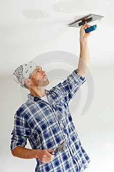 Young man plastering damages in ceiling with trowel prior to painting