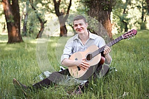 Young man playing guitar in the forest, sit on the grass, summer nature, bright sunlight, shadows and green leaves, romantic feeli