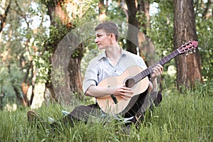 Young man playing guitar in the forest, sit on the grass, summer nature, bright sunlight, shadows and green leaves, romantic feeli