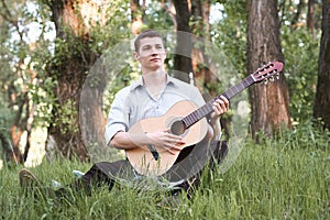 Young man playing guitar in the forest, sit on the grass, summer nature, bright sunlight, shadows and green leaves, romantic feeli