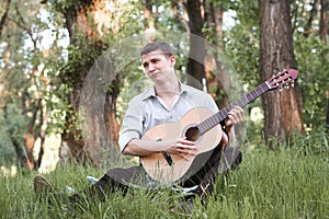 Young man playing guitar in the forest, sit on the grass, summer nature, bright sunlight, shadows and green leaves, romantic feeli