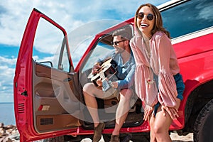 young man playing guitar near car with cheerful girlfriend