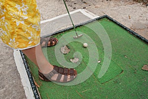 Young man playing minigolf on old, worn hotel resort green field, closeup to his feet in sandals, metal club and ball