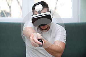 Young man playing a shooting video game with virtual reality headset and a joystick sitting on sofa