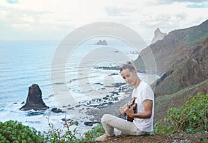 Young man Playing Ukulele on a Grassy Cliff Overlooking the Ocean Waves Crashing Against Rocky Shoreline