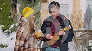 Young man playing ukulele singing serenade for smiling woman holding coffee tea cup outdoors on sunny winter day