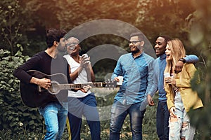 A young man plays the guitar in a multiethnic company of five people. Friends having fun in a park during sunset. Impromptu jam photo