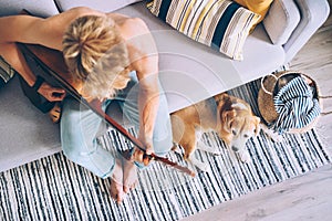 Young man plays on guitar sitting on sofa and beagle dog listen