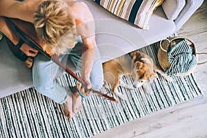 Young man plays on guitar sitting on sofa in cozy home atmosphere. Beagle dog lies near on the floor