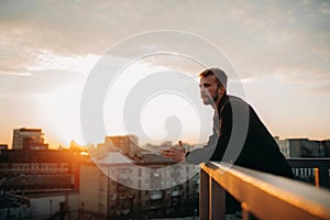 Young man is pondering on terrace of roof against background of cityscape at sunset.