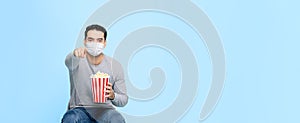 Young man with popcorn protect himself by wearing medical face mask while enjoying watching movie  on blue banner