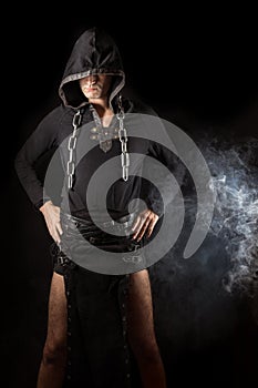 Young man posing in hooded gothic clothes with chain around neck next to smoke