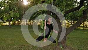 A young man practices aerial yoga in a hammock hanging from a tree in the park.