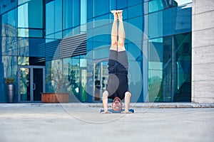 Young man practices headstand yoga asana Sirsasana pose outdoors against the background of a modern city