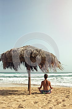 Young man practicing yoga at the beach. He sits in pose of lotus under big cane sun umbrella and looks at the sea