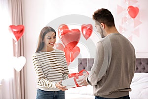 Young man presenting gift to his girlfriend in bedroom decorated with heart balloons. Valentine`s day celebration