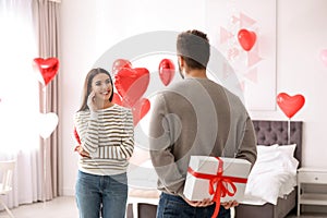 Young man presenting gift to his girlfriend in bedroom decorated with heart balloons. Valentine`s day celebration