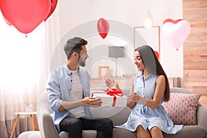 Young man presenting gift to his girlfriend in room decorated with heart shaped balloons. Valentine`s day celebration