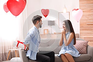 Young man presenting gift to his girlfriend in room decorated with heart shaped balloons. Valentine`s day celebration