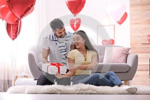 Young man presenting gift to his girlfriend in room decorated with heart shaped balloons. Valentine`s day celebration