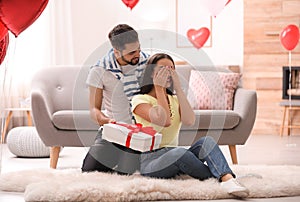 Young man presenting gift to his girlfriend in room decorated with heart shaped balloons. Valentine`s day celebration