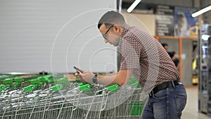 A young man prints a message on the screen of his smartphone, leaning on the shopping cart in the supermarket. Using