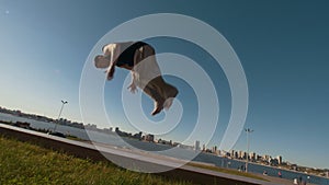 Young man professional acrobat performs a flip on a cliff overlooking the river and cityscape at sunset