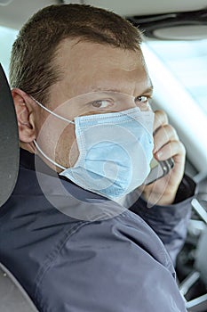 A young man in a protective mask is sitting in a left-hand drive car and calls on his mobile phone. Taxi driver. Coronavirus