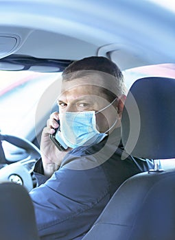 A young man in a protective mask is sitting in a right-hand drive car and calls on his mobile phone. Taxi driver. Coronavirus