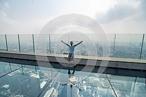 Young man with raised haands on a glass floor on the roof of a King Power Mahanakhon building