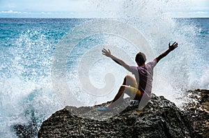 Young man with raised hands sitting on the rock with the sea waves