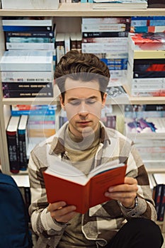 Young man reading book intently in library surrounded by literature