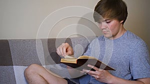 Young man is reading book intently sitting on sofa. Concept. Student reads classics with strained look. Man reads old