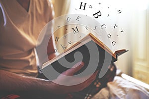 Young man reading book with letters flying over it on bed at home, closeup