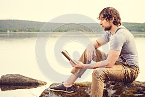 Young Man reading book outdoor with scandinavian lake on background