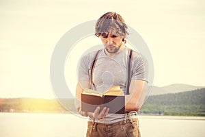 Young Man reading book outdoor with scandinavian lake on background