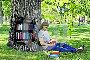 Young man reading a book sitting on the green grass leaning on a tree in the Park.