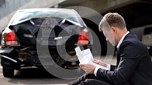 Young man reading insurance document sitting road on broken car background